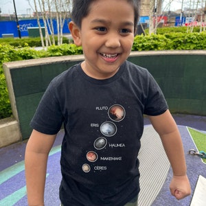 Puede incluir: Un niño pequeño con una camiseta negra con un gráfico de los planetas enanos Plutón, Eris, Haumea, Makemake y Ceres. La camiseta tiene los nombres de los planetas impresos debajo de cada imagen. El niño sonríe y mira hacia la derecha. Lleva pantalones de camuflaje.