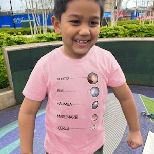Puede incluir: Un niño pequeño con una camiseta rosa con un gráfico de los planetas enanos Plutón, Eris, Haumea, Makemake y Ceres. El gráfico incluye los nombres de los planetas enanos y una pequeña imagen de cada uno.