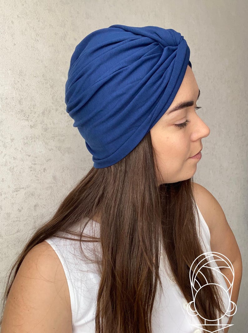 May include: A solid dark blue turban hat featuring a gathered, knotted design at the front. The fabric is smooth and opaque, creating soft folds and pleats that wrap around the head. The hat provides full coverage with a snug, fitted silhouette.