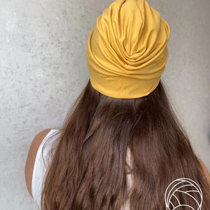 Pode incluir: Cobertura de cabe&ccedil;a estilo turbante amarelo mostarda. O tecido &eacute; franzido e drapeado, criando um efeito texturizado e em camadas. A cobertura da cabe&ccedil;a fica sobre cabelos longos e castanhos. A pessoa est&aacute; usando uma regata branca.