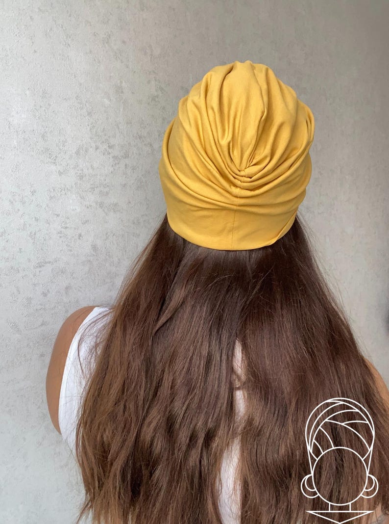 Pode incluir: Cobertura de cabe&ccedil;a estilo turbante amarelo mostarda. O tecido &eacute; franzido e drapeado, criando um efeito texturizado e em camadas. A cobertura da cabe&ccedil;a fica sobre cabelos longos e castanhos. A pessoa est&aacute; usando uma regata branca.