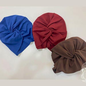 May include: Three turbans in solid colors: one bright blue, one deep burgundy red, and one dark brown. Each turban features a central twisted knot detail and gathered fabric construction, creating a soft, rounded shape with visible folds.
