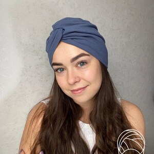 May include: A dusty blue turban-style head wrap featuring a gathered, knotted front detail. The fabric is smooth and opaque, designed to fit snugly around the head with soft folds that create a layered appearance across the crown and sides.