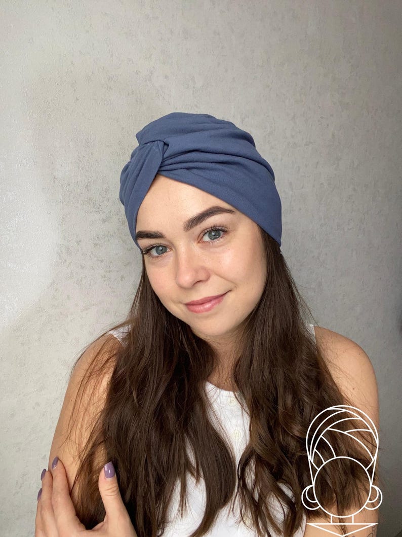 May include: A dusty blue turban-style head wrap featuring a gathered, knotted front detail. The fabric is smooth and opaque, designed to fit snugly around the head with soft folds that create a layered appearance across the crown and sides.