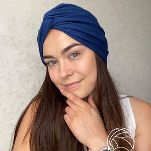 May include: A solid dark blue turban-style head covering featuring a gathered, twisted knot detail at the center front. The fabric is smooth and opaque, designed to fit snugly around the head with a rounded silhouette and soft, draped folds.