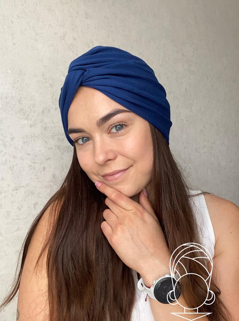 May include: A solid dark blue turban-style head covering featuring a gathered, twisted knot detail at the center front. The fabric is smooth and opaque, designed to fit snugly around the head with a rounded silhouette and soft, draped folds.