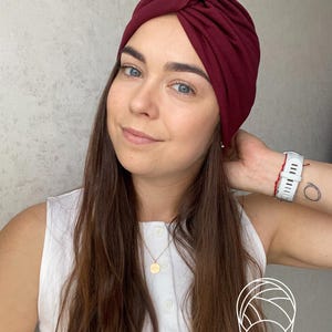 May include: A deep burgundy turban-style head wrap featuring a central twisted knot detail at the forehead. The fabric is smooth and solid-colored, designed to fit snugly around the head, providing full coverage with a gathered, layered appearance.