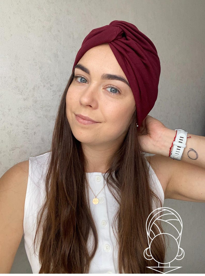 May include: A deep burgundy turban-style head wrap featuring a central twisted knot detail at the forehead. The fabric is smooth and solid-colored, designed to fit snugly around the head, providing full coverage with a gathered, layered appearance.