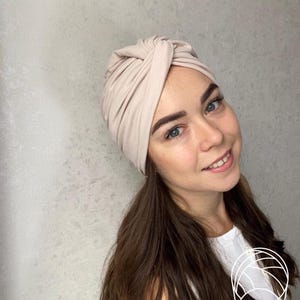 Pode incluir: Um turbante bege. O tecido &eacute; macio e parece ser um material de malha. O turbante &eacute; amarrado no topo e &eacute; usado na cabe&ccedil;a. A pessoa que usa o turbante tem cabelo castanho comprido e est&aacute; sorrindo.