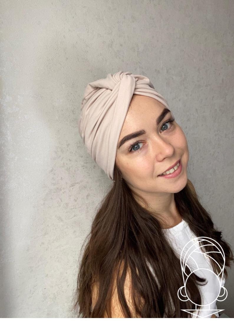 Pode incluir: Um turbante bege. O tecido &eacute; macio e parece ser um material de malha. O turbante &eacute; amarrado no topo e &eacute; usado na cabe&ccedil;a. A pessoa que usa o turbante tem cabelo castanho comprido e est&aacute; sorrindo.