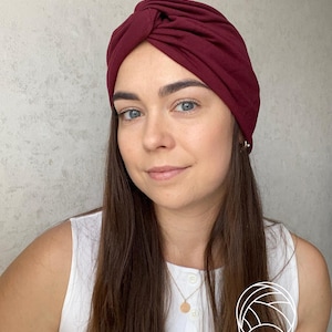 May include: A deep burgundy turban-style head wrap featuring a central knotted detail at the front. The fabric is smooth and opaque, covering the crown of the head completely. A small white line-art icon of a head wrap is visible in the corner.