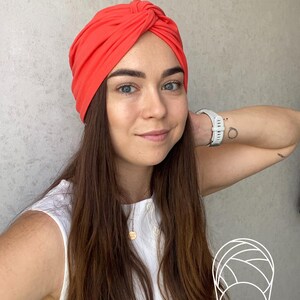 Pode incluir: Um turbante vibrante de cor coral. O adorno de cabe&ccedil;a &eacute; amarrado no topo e feito de um tecido macio. A mulher na imagem est&aacute; usando um top branco e um rel&oacute;gio branco.