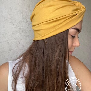 Pode incluir: Enrolamento de cabe&ccedil;a estilo turbante amarelo mostarda. O tecido &eacute; macio e franzido, com um detalhe de n&oacute; na parte superior. O adorno de cabe&ccedil;a &eacute; usado por uma pessoa com cabelo castanho comprido e uma regata branca.