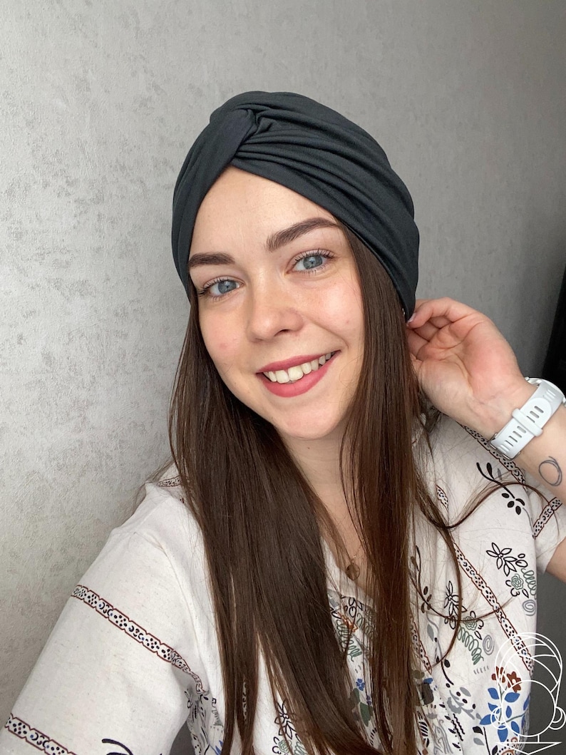 Pode incluir: Um turbante cinza escuro. O tecido &eacute; macio e parece drapeado. O turbante &eacute; usado por uma pessoa com cabelo castanho comprido e uma camisa branca estampada. A pessoa est&aacute; sorrindo.