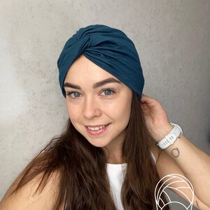 May include: A dark teal turban-style head wrap featuring a central twisted knot detail at the front. The fabric is smooth and fitted, covering the crown of the head securely. A small white line-art icon of a turban is visible in the corner.