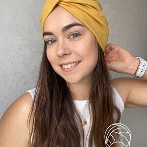 Pode incluir: Faixa de cabe&ccedil;a estilo turbante amarelo mostarda. O tecido &eacute; franzido e torcido na parte superior, criando um acess&oacute;rio elegante e confort&aacute;vel. A faixa de cabe&ccedil;a &eacute; usada por uma pessoa com cabelo castanho comprido e um top branco.