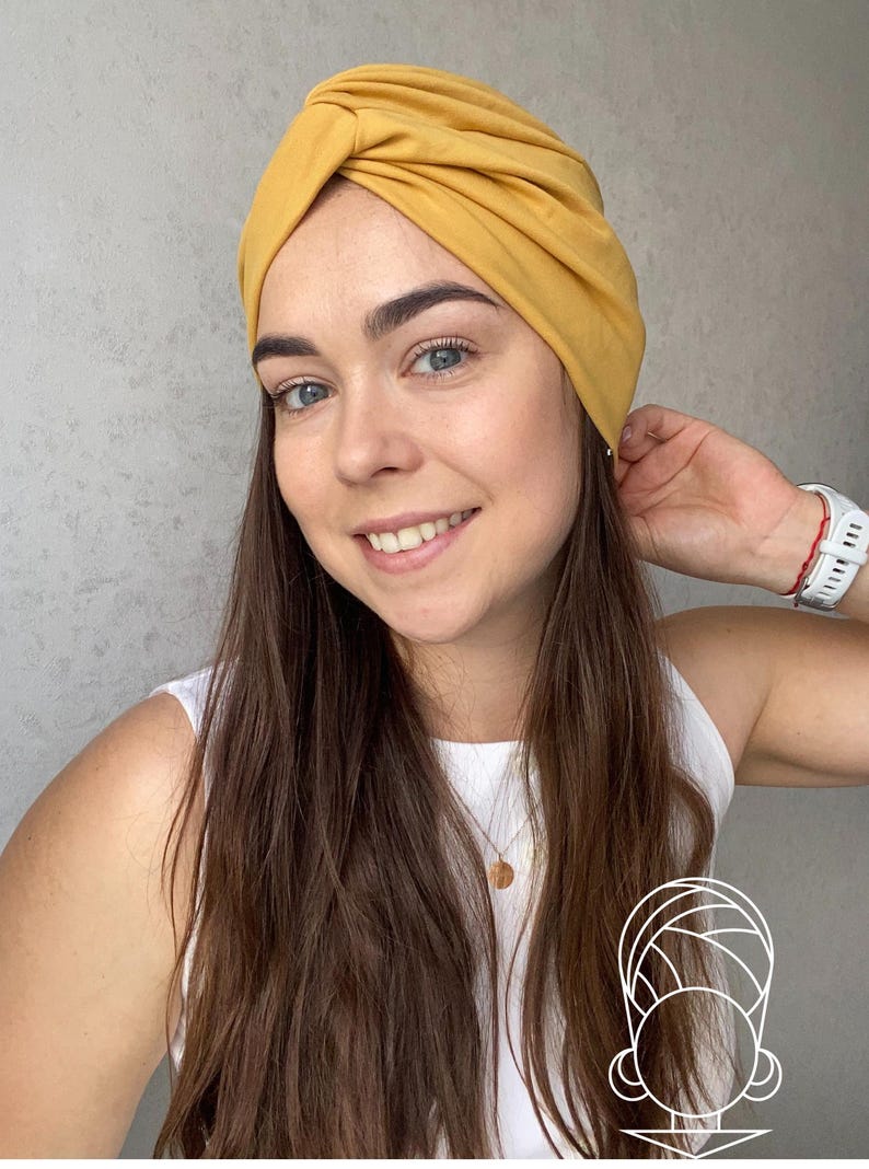 Pode incluir: Faixa de cabe&ccedil;a estilo turbante amarelo mostarda. O tecido &eacute; franzido e torcido na parte superior, criando um acess&oacute;rio elegante e confort&aacute;vel. A faixa de cabe&ccedil;a &eacute; usada por uma pessoa com cabelo castanho comprido e um top branco.