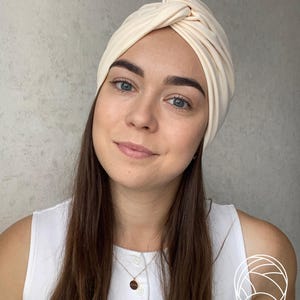 Pode incluir: Um turbante creme com um design de n&oacute; torcido. A cobertura da cabe&ccedil;a &eacute; feita de tecido macio. O turbante &eacute; um acess&oacute;rio elegante. A pessoa est&aacute; usando um top branco e um colar de ouro.