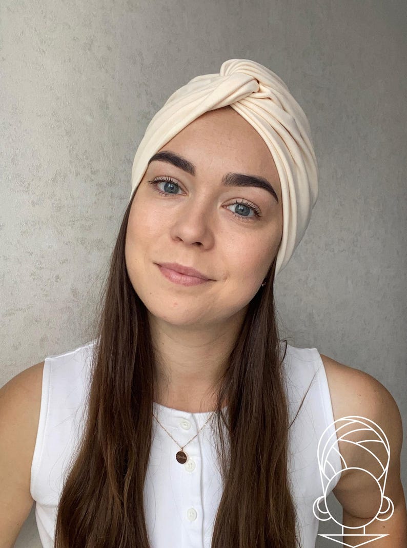Pode incluir: Um turbante creme com um design de n&oacute; torcido. A cobertura da cabe&ccedil;a &eacute; feita de tecido macio. O turbante &eacute; um acess&oacute;rio elegante. A pessoa est&aacute; usando um top branco e um colar de ouro.