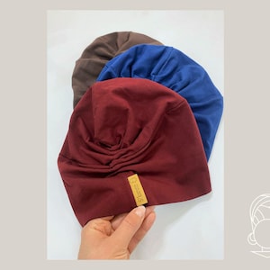 May include: Three soft fabric turbans in burgundy, royal blue, and brown. Each turban features a gathered, pleated design at the front and a small rectangular tan label with the text S'tarute B stitched onto the lower edge of the burgundy piece.