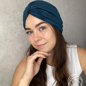 May include: A dark teal turban-style head wrap featuring a gathered, twisted knot detail at the center front. The fabric is smooth and opaque, covering the crown of the head completely with a snug fit that contours around the forehead and ears.