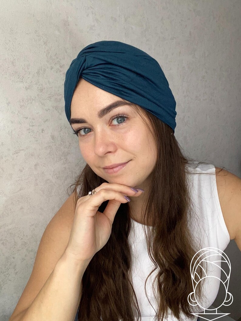 May include: A dark teal turban-style head wrap featuring a gathered, twisted knot detail at the center front. The fabric is smooth and opaque, covering the crown of the head completely with a snug fit that contours around the forehead and ears.