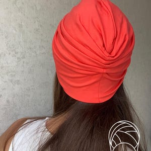 Pode incluir: Um turbante vibrante na cor coral. O adorno de cabe&ccedil;a &eacute; feito de tecido macio e &eacute; franzido na parte superior. O turbante &eacute; mostrado por tr&aacute;s, com cabelos castanhos longos e uma parte superior branca vis&iacute;vel.