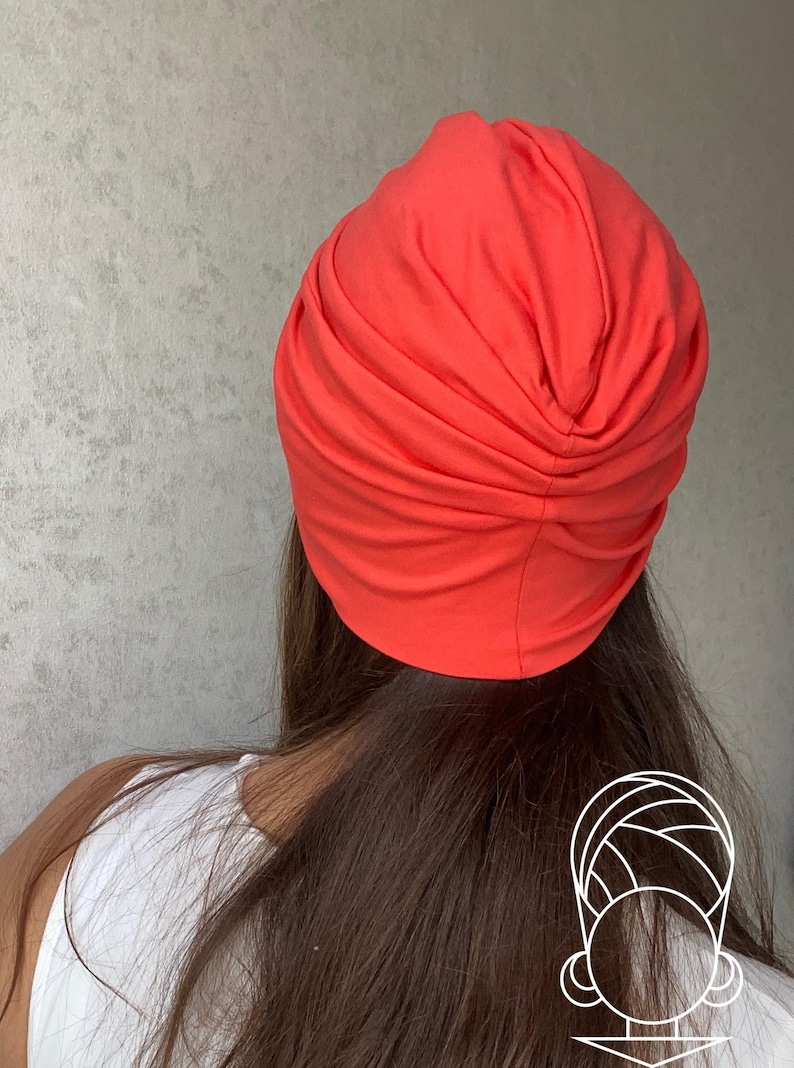 Pode incluir: Um turbante vibrante na cor coral. O adorno de cabe&ccedil;a &eacute; feito de tecido macio e &eacute; franzido na parte superior. O turbante &eacute; mostrado por tr&aacute;s, com cabelos castanhos longos e uma parte superior branca vis&iacute;vel.