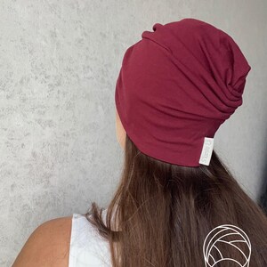 May include: A deep burgundy beanie hat with a slouchy, gathered design at the back. The hat features a small white fabric tag on the lower edge with the text S'karite printed in black. The material appears to be a soft, smooth jersey knit fabric.