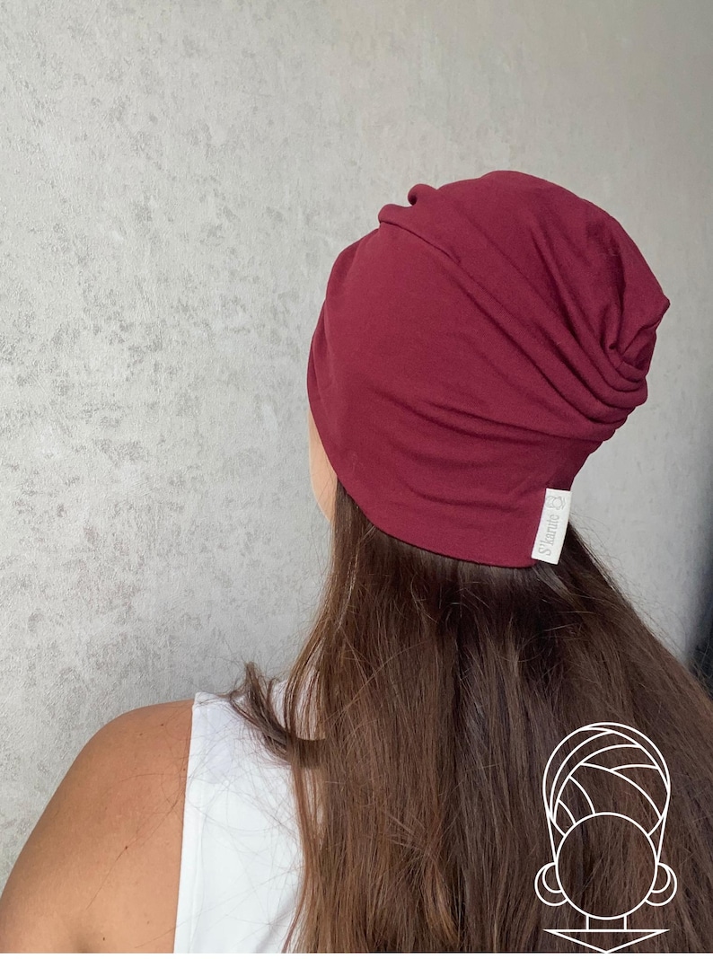 May include: A deep burgundy beanie hat with a slouchy, gathered design at the back. The hat features a small white fabric tag on the lower edge with the text S'karite printed in black. The material appears to be a soft, smooth jersey knit fabric.