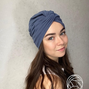 May include: A dusty blue turban-style head wrap featuring a gathered, knotted front detail. The fabric is smooth and matte, designed to fit snugly around the head with soft folds that create a textured, layered appearance across the crown.
