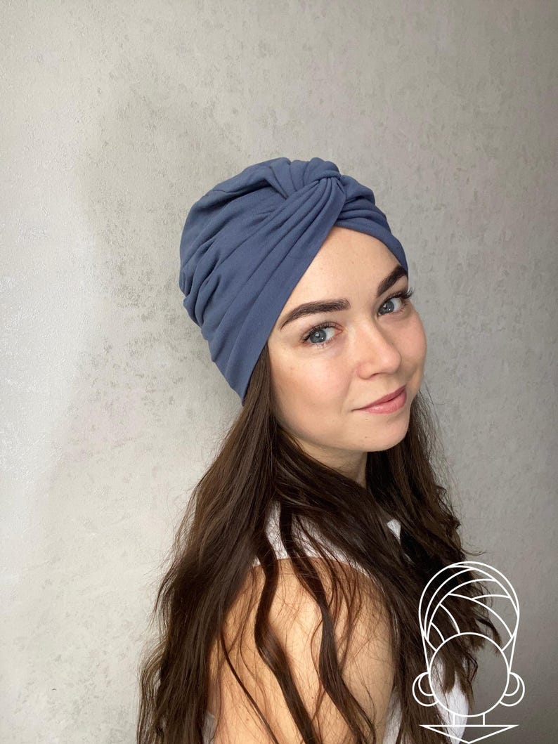 May include: A dusty blue turban-style head wrap featuring a gathered, knotted front detail. The fabric is smooth and matte, designed to fit snugly around the head with soft folds that create a textured, layered appearance across the crown.