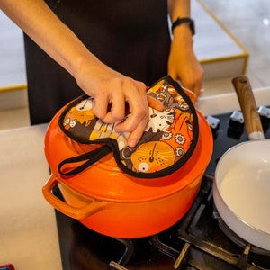 May include: A bright orange pot with a black handle and a colourful pot holder with a cat design on top. The pot is on a black hob.