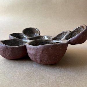 Può includere: Un piatto in ceramica a più scomparti con un esterno ruvido color marrone rossastro scuro e un interno smaltato liscio e lucido di colore chiaro. L'oggetto presenta diverse ciotole arrotondate interconnesse in un grappolo organico.