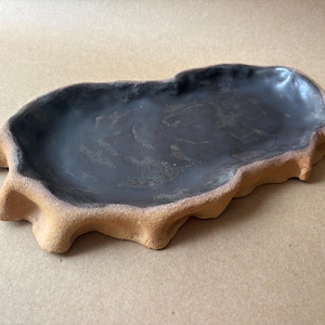 May include: An oblong ceramic tray featuring a dark, glossy black interior basin with a smooth finish. The exterior rim has a textured, matte tan surface with an irregular, organic shape that mimics the appearance of natural rock formations.