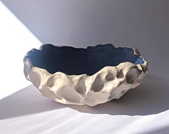 May include: A ceramic bowl with a highly textured, off-white exterior featuring deep, irregular indentations and a smooth, dark navy blue interior. The rim of the bowl is uneven and organic in shape, contrasting with the dark, matte-finished basin.