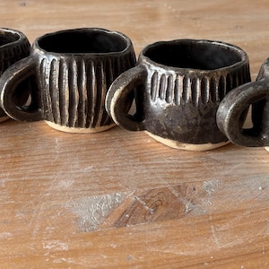 Può includere: Un set di quattro piccole tazze in ceramica marrone scuro con corpi arrotondati e manici spessi. Ogni tazza presenta una texture unica intagliata a mano sull'esterno, tra cui linee verticali, tratteggi incrociati e trattini, con base beige.
