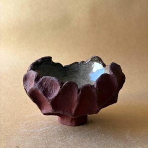 Può includere: Una piccola ciotola in ceramica formata a mano con un esterno scuro, opaco e bruno-rossastro caratterizzato da un bordo smerlato simile a un petalo. L'interno della ciotola è rifinito con uno smalto grigio chiaro liscio e lucido che contrasta con la superficie esterna ruvida.