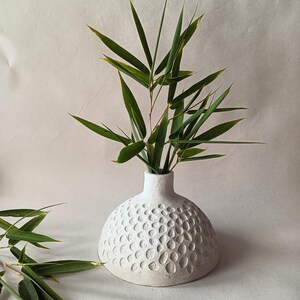 Pode incluir: Vaso de cerâmica branco com uma superfície texturizada e pontilhada. O vaso está cheio de ramos verdes com folhas.