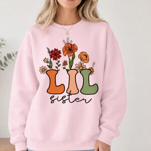 Può includere: Una felpa girocollo rosa chiaro con una stampa grafica di fiori colorati che emergono dalle lettere LIL in arancione, pesca e verde salvia, con la parola sister scritta in corsivo nero sotto il disegno floreale.
