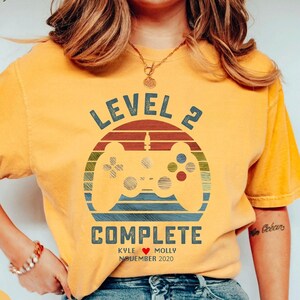 May include: A mustard yellow t-shirt with a retro-style graphic of a video game controller and the text "Level 2 Complete" with the names "Kyle" and "Molly" and the date "November 2020".