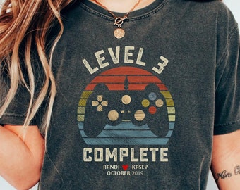 May include: A dark gray t-shirt with a vintage-style graphic of a video game controller and the text "Level 3 Complete". The graphic is set against a sunset background with red, orange, and blue colors. The text "Randi Kasey October 2019" is printed below the graphic.