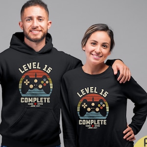 May include: A black hoodie and sweatshirt with a retro-style graphic of a video game controller and the text "Level 15 Complete". The graphic has a sunset background with orange, yellow, and red colors. The text "Level 15" is in white and the text "Complete" is in yellow. The text "Noah & Emma June 2017" is in white below the graphic.