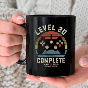 May include: Black ceramic mug featuring the text "LEVEL 20 COMPLETE" and a retro video game controller design. The mug includes the names Pamela and Steven, and the date October 2002. The mug is held in a person's hands.