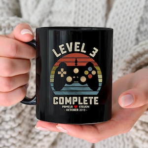 May include: Black ceramic mug with a retro-style video game controller graphic and the text "Level 3 Complete" with a heart and the names "Pamela & Steven" and the date "October 2019".