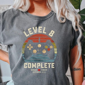 May include: A grey t-shirt with a vintage-style graphic of a video game controller and the text "Level 8 Complete". The graphic also includes the text "Randi & Kasey October 2014".
