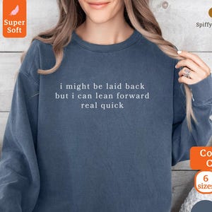 Puede incluir: Una sudadera azul denim con el texto "i might be laid back but i can lean forward real quick" en blanco. La sudadera tiene cuello redondo y mangas largas. La imagen también muestra las palabras "Comfort Colors", "6 tallas" y "8 colores".