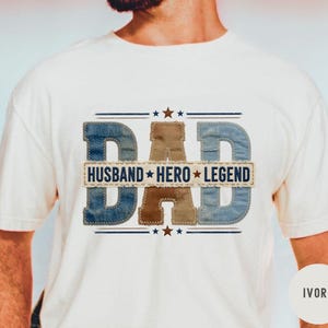 May include: Ivory t-shirt featuring the word "DAD" in denim-style lettering, with "HUSBAND HERO LEGEND" below. The design incorporates stars and is a thoughtful gift. The t-shirt is a cream colour.