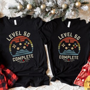 May include: Two black t-shirts with a retro video game controller graphic and the text "Level 50 Complete". The shirts are personalized with the names "Pamela" and "Steuen" and the date "October 1972".