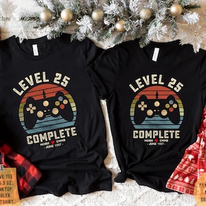 May include: A black t-shirt with a retro-style video game controller graphic and the text "Level 25 Complete". The graphic is set against a sunset background with a red, orange, and yellow color scheme. The text "Noah Emma June 1997" is printed below the graphic.